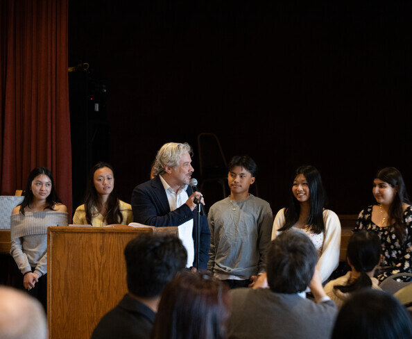 a man speaking into a microphone with a group of people in the background
